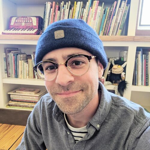 PhilStead-500x500 | The Children's Book Review Smiling man with blue knit beanie and round glasses in a cozy library, bookshelves behind him at home or school.