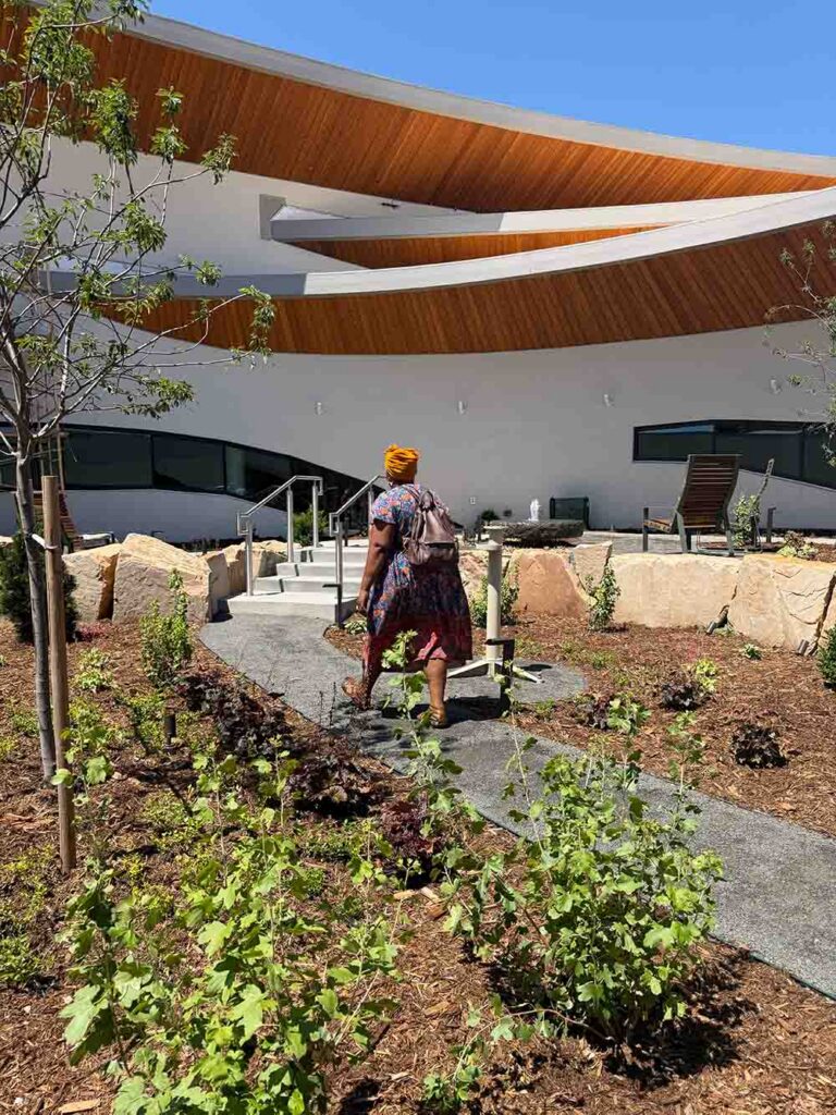 Anything Library Center Garden | The Children's Book Review Woman with an orange head wrap walks along a concrete path in a desert-style garden toward a modern white building with curved wooden roof canopies in bright sun.
