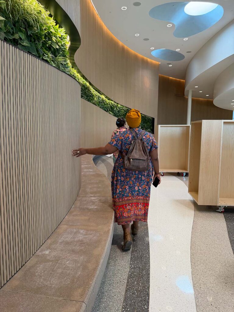 Inside Anythink Library | The Children's Book Review Person wearing a colorful floral dress and mustard head wrap walks down a curved indoor hallway with wooden walls and greenery above, carrying a backpack and phone