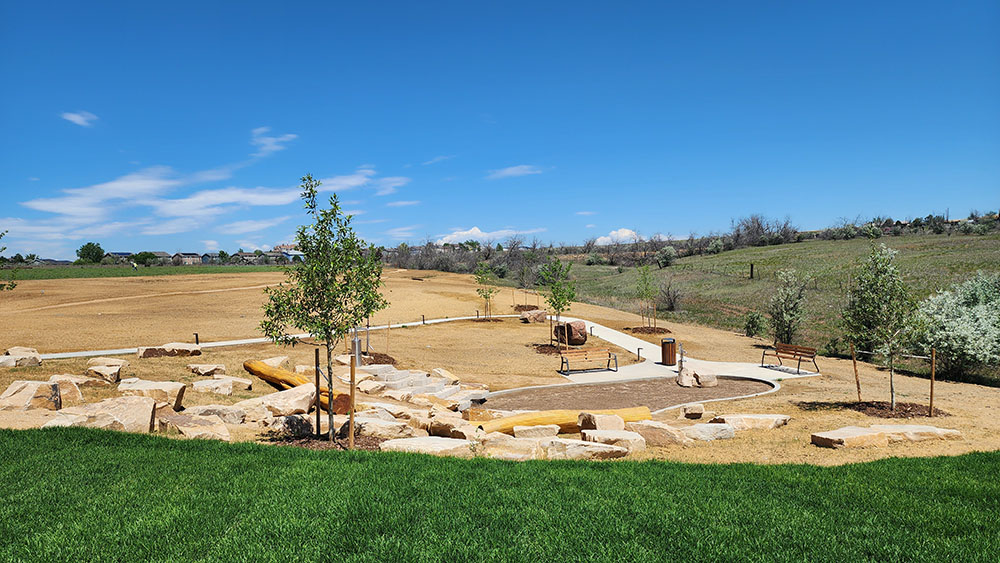 Mud Pit at Anythink | The Children's Book Review Wide view of a park with a winding concrete path, wooden benches, and a rock landscape under a clear blue sky.