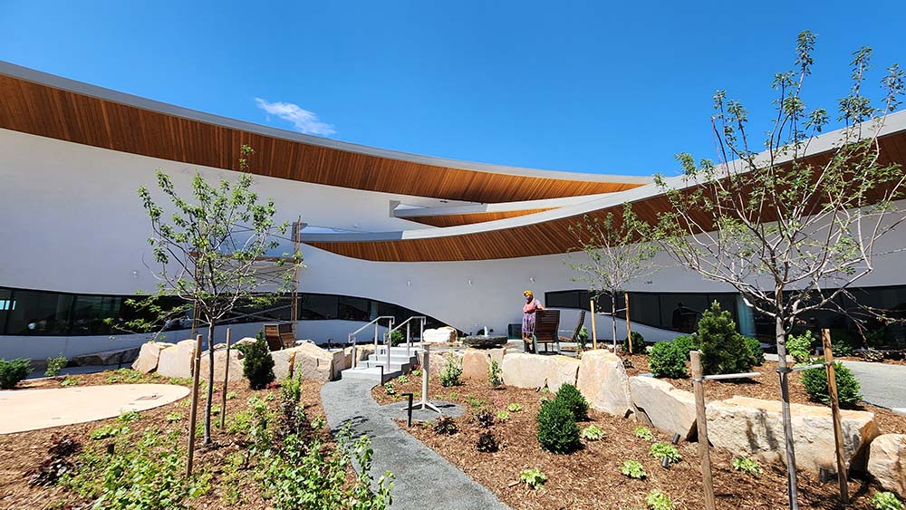 Three Rooflines Anythink Library | The Children's Book Review Contemporary courtyard with curved wooden canopies over a white building and landscaped garden path, a person standing near benches in the background.